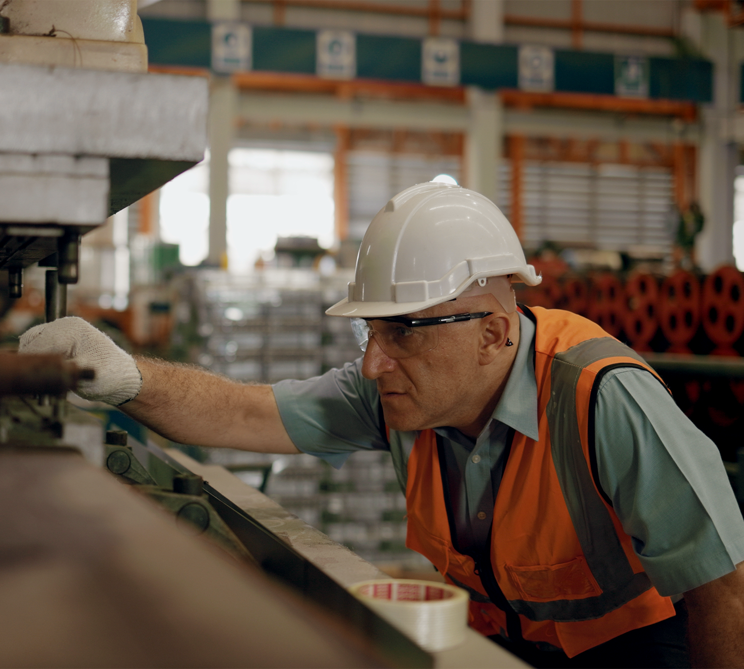 A worker in a hard hat and safety vest inspecting material on a factory line